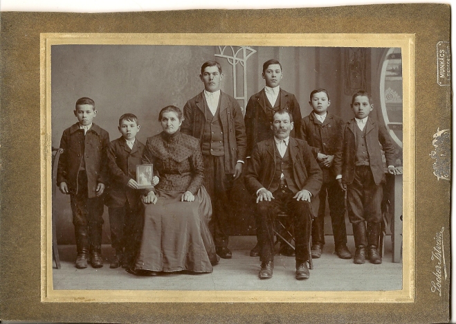 An old family photo with 6 boys standing behind a man and woman who are sitting. One of the boys holds a photograph. 